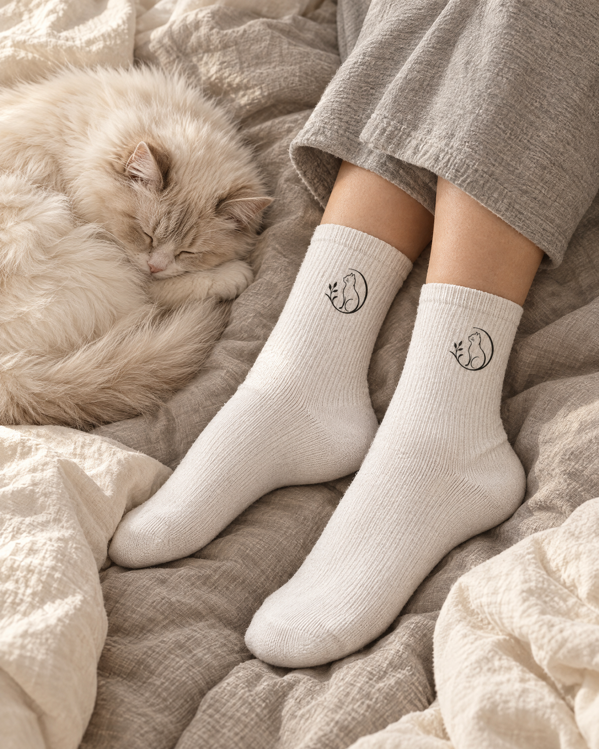 Weiße Mr. Fluffel Socken mit Katzen-Logo, getragen auf dem Bett neben einer schlafenden Katze